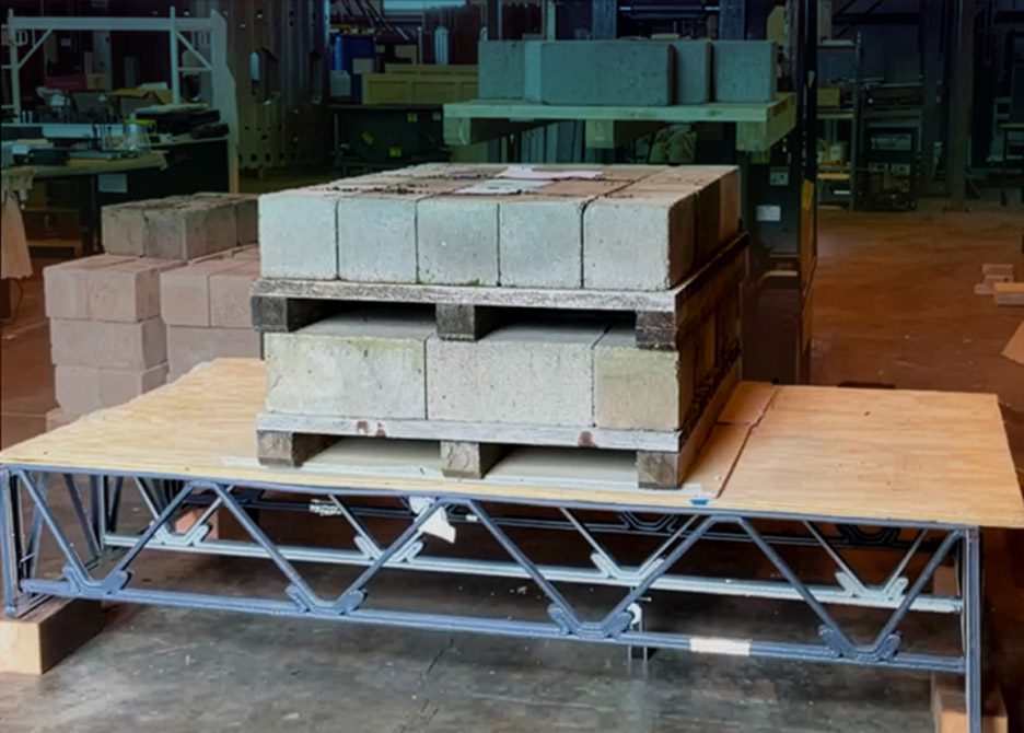 Photo of a floor truss being tested with concrete blocks piled on top of it. The truss is made of a plastic composite 3D-printed into a zigzag pattern below a plywood top